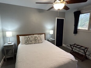 Un dormitorio con una cama, dos lámparas de noche, un ventilador de techo, una ventana con cortinas y un banco de madera.