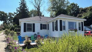 Ein eingeschossiges Mobilheim mit Satteldach, weißer Außenverkleidung und schwarzen Fensterläden. Es besitzt eine kleine Veranda mit bunten Adirondack-Stühlen und ein Blumenbeet mit violetten Blüten.