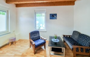 A cozy living room with a wooden ceiling, a blue patterned armchair, a matching sofa, a glass coffee table with a plant, and a window with a view of greenery.