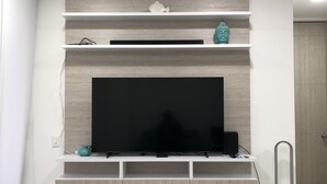 Uma sala de estar moderna com uma televisão de tela plana presa a uma parede de madeira, uma prateleira pequena com objetos decorativos e um ventilador de chão.