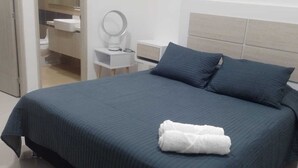 Cama bem arrumada com colcha azul escura e dois travesseiros, mesa de cabeceira branca com espelho redondo e um criado-mudo branco.