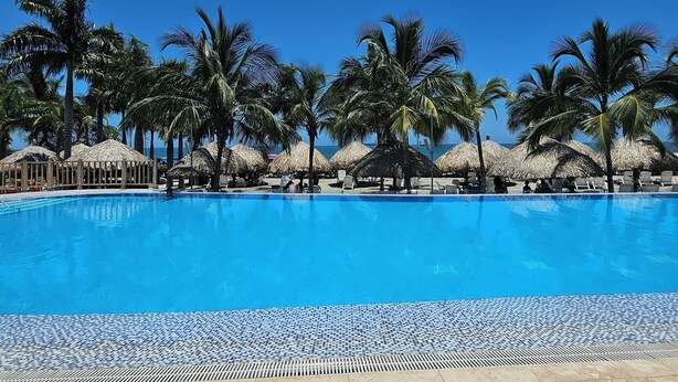 Piscina azul límpida rodeada por palmeiras e guarda-sóis de palha, com um deck de azulejos em primeiro plano.