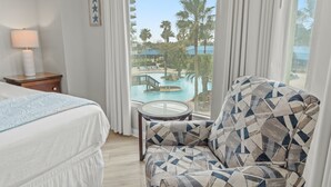 A hotel room with a bed, a patterned armchair, a small table, and a view of a pool and palm trees. - 2317 Gorgeous 3 Bdrm ~ Lagoon Pool ~ Hot Tub ~ Restaurant ~ Bar ~ Book Sept 27 (Destin)