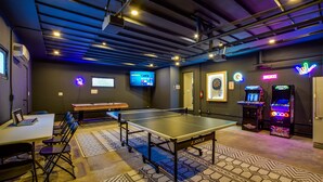 A room with a ping pong table, chairs, a dartboard, and arcade machines.