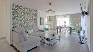 A modern living room with a sofa, coffee table, and a patterned wall. There is a dining area with chairs and a table, and a kitchen in the background.