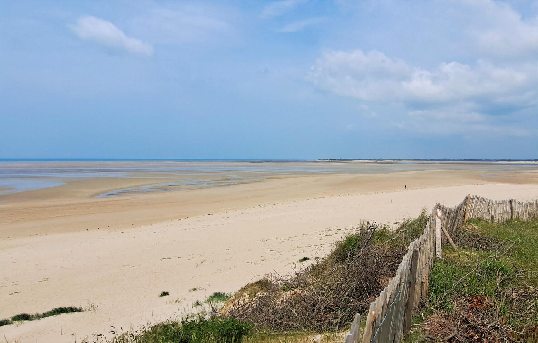 A wide sandy beach with gentle waves, a clear sky, and a wooden fence in the foreground.