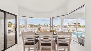 Un comedor con una mesa dispuesta para cuatro personas, amplias ventanas que ofrecen vistas a una piscina y una zona residencial.