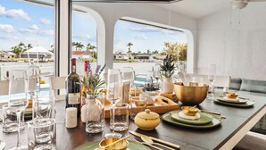 Una mesa de comedor dispuesta con diferentes vasos, una botella y adornos en una habitación con ventanas amplias y vistas a una zona residencial.