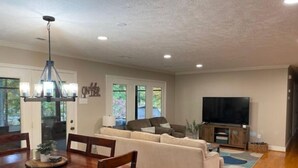 A spacious living room with a large sofa, a dining table with chairs, a chandelier, and a flat-screen TV. - License To Chill - Lakefront/Screened in Porch (Bumpass)