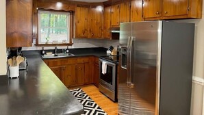 A kitchen with wooden cabinets, a stainless steel refrigerator, a stove, and a window with a view of trees. - License To Chill - Lakefront/Screened in Porch (Bumpass)