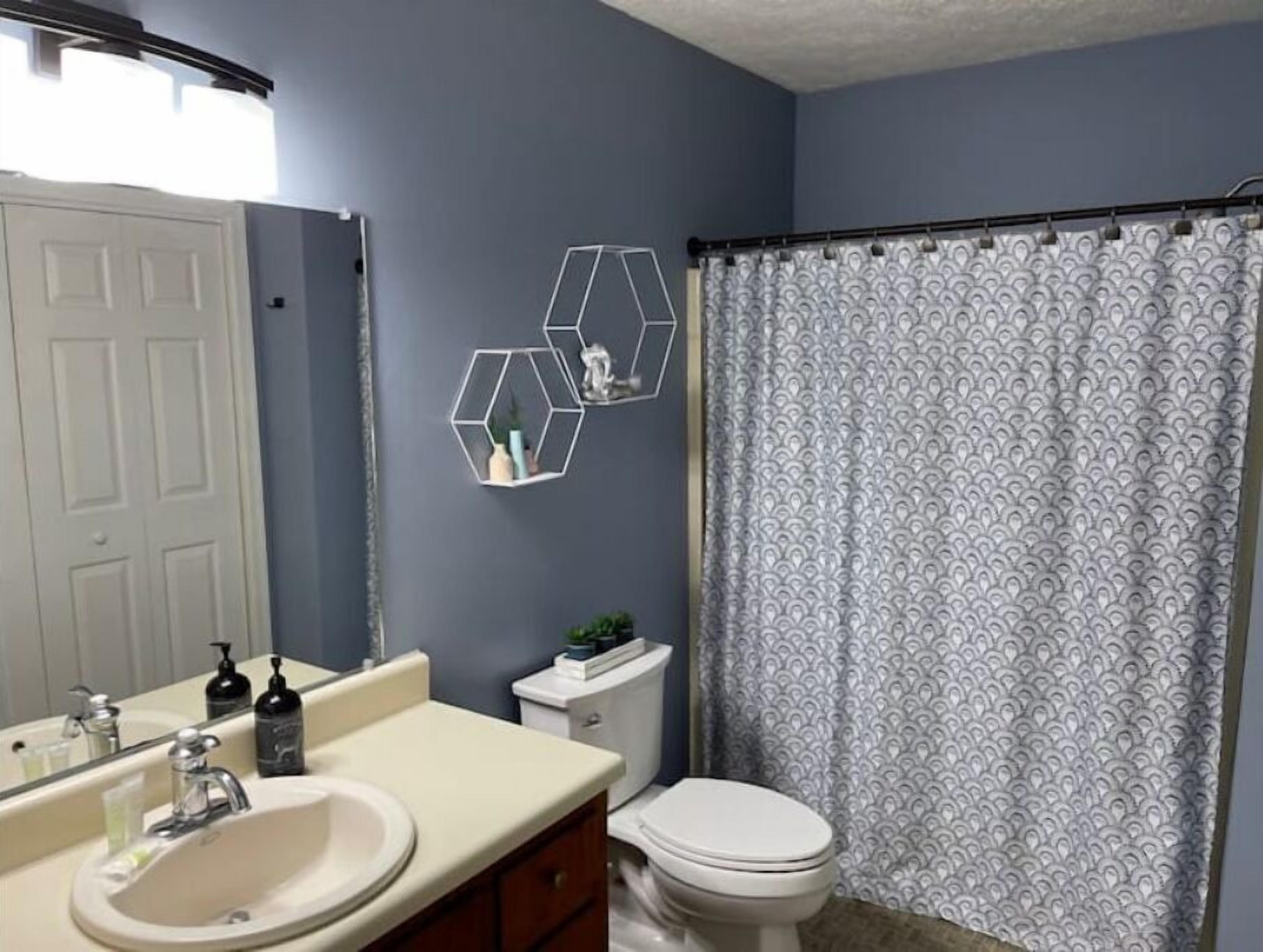 Une salle de bain avec un lavabo blanc, un miroir et des toilettes. On y trouve deux décorations murales hexagonales et un rideau de douche à motifs.