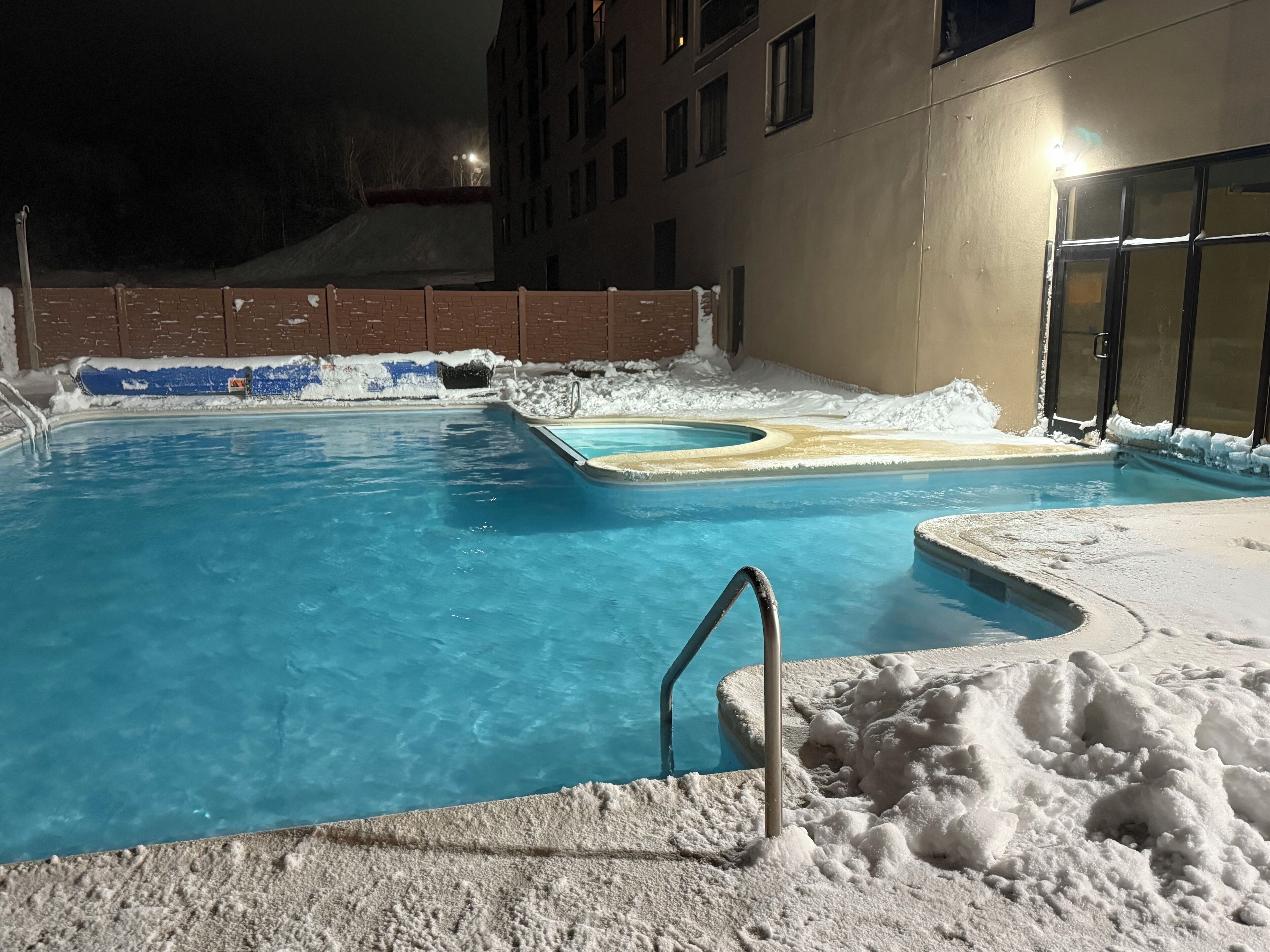 A swimming pool surrounded by snow, with a brick wall and a building in the background.