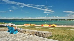 Zona de playa con un paseo pavimentado, dos sillas azules y vista al mar con barcos.