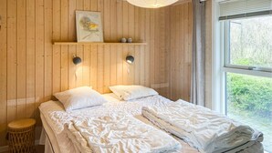 A bedroom with a wooden wall, a bed with white bedding, two wall-mounted lamps, a shelf with decorative items, and a window with a view of greenery. - Awesome home in Sydals with kitchen (Sydals)