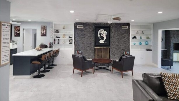 A modern living room with a dining area, featuring a central coffee table, two armchairs, and a sofa. There is a kitchen area with a white countertop and bar stools. The room has a stone accent wall and a ceiling fan.
