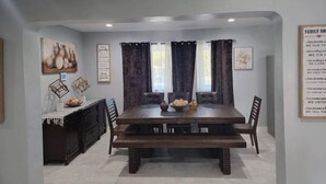 A dining room with a large wooden table, dark curtains, and a wall-mounted sign with family values.