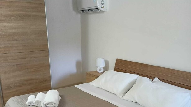 A hotel room with a bed, pillows, and rolled towels on the bed. There is a nightstand with a lamp and a wall-mounted air conditioning unit. - Cozy Little House in town La Ficurinia 🌵 (Siracusa)