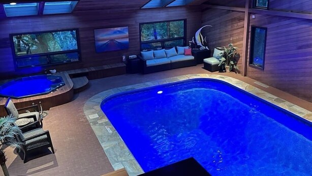 Een modern binnenzwembad met een houten vloer, een jacuzzi en een zithoek met een bank en stoelen.