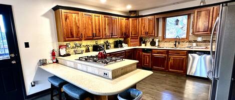 Une cuisine moderne avec des meubles en bois, un îlot central doté de tabourets, un réfrigérateur en acier inoxydable et une fenêtre offrant une vue agréable.