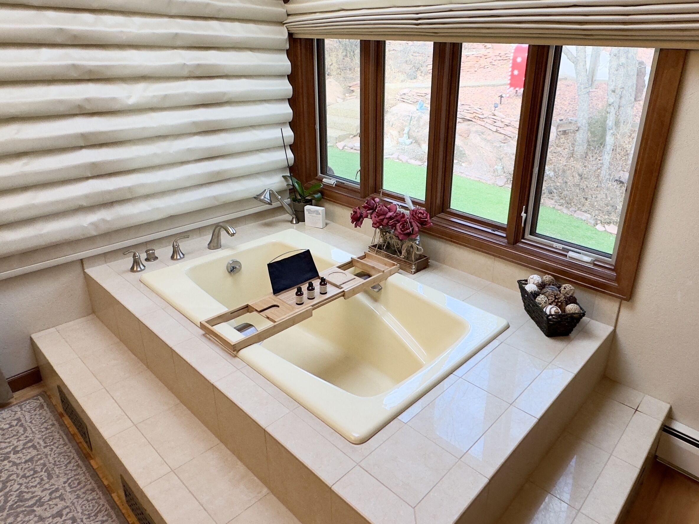 Une salle de bain avec une grande baignoire, une fenêtre avec des stores et une petite plante posée sur le rebord de la fenêtre.