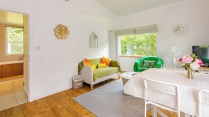 Una habitación con un sofá verde, una mesa blanca, un televisor y una ventana con vistas a árboles.