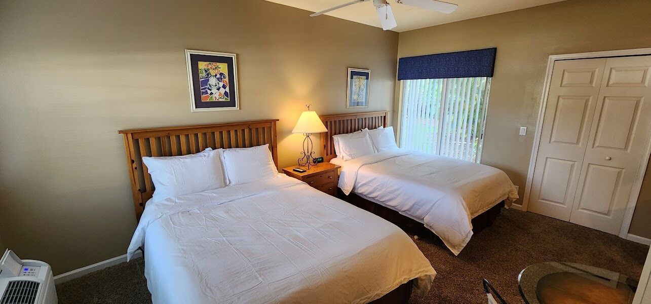 A hotel room with two beds, a bedside table with a lamp, a ceiling fan, and a door.