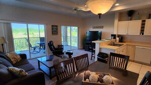 A spacious living area with a sofa, coffee table, and chairs, a balcony with a view, and a kitchen with white cabinets and appliances. - Right Direction Rentals (Port Saint Lucie)
