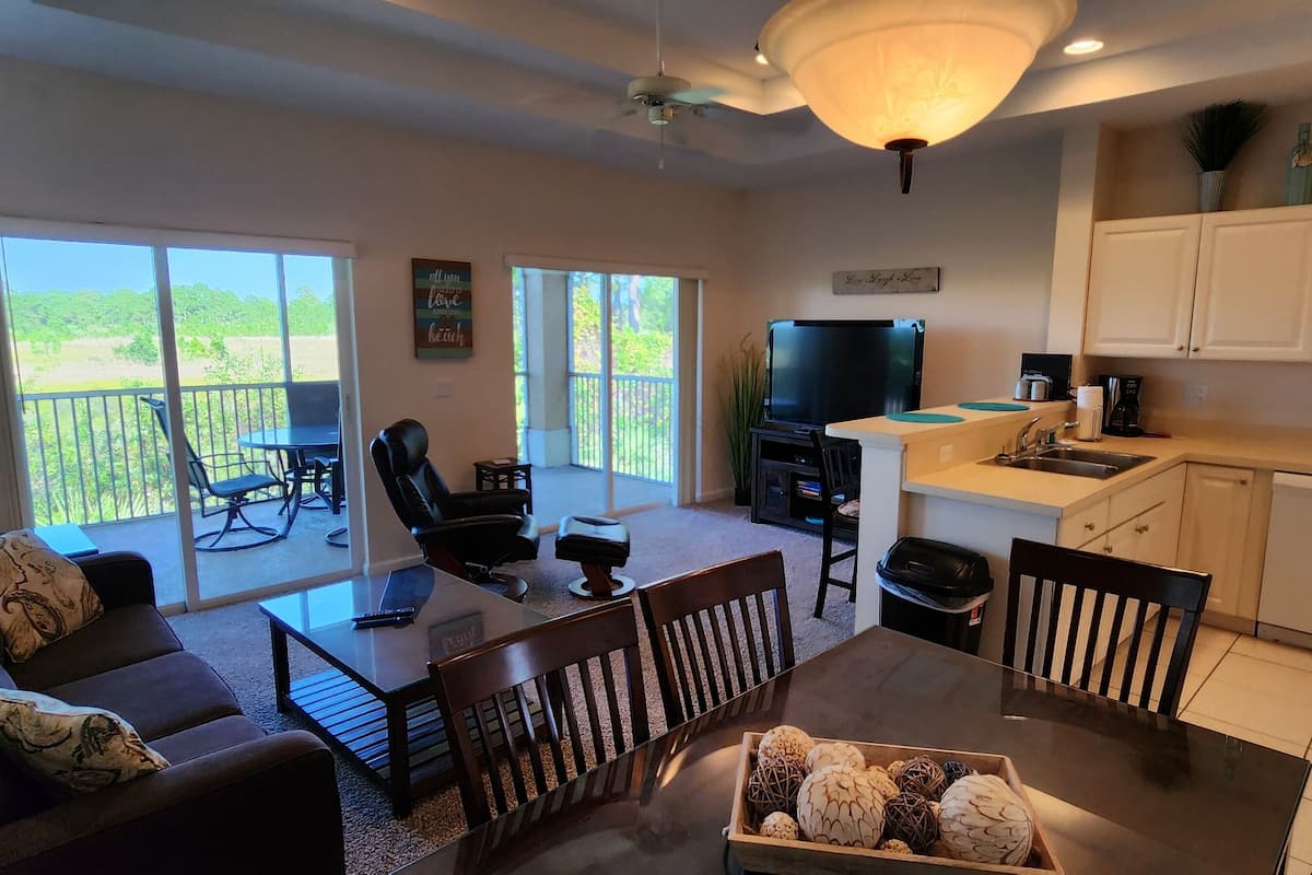 A spacious living area with a sofa, coffee table, and chairs, a balcony with a view, and a kitchen with white cabinets and appliances.
