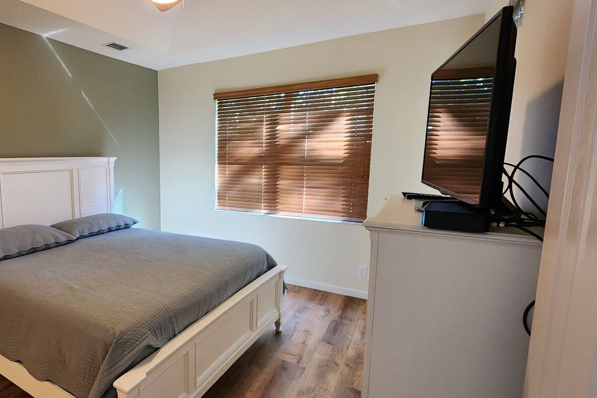 A bedroom with a bed, a nightstand, a dresser, a window with blinds, and a TV.