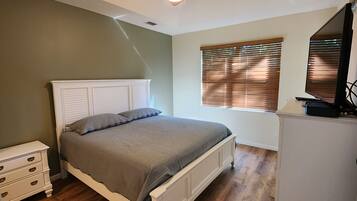 A bedroom with a bed, a nightstand, a dresser, a window with blinds, and a TV.