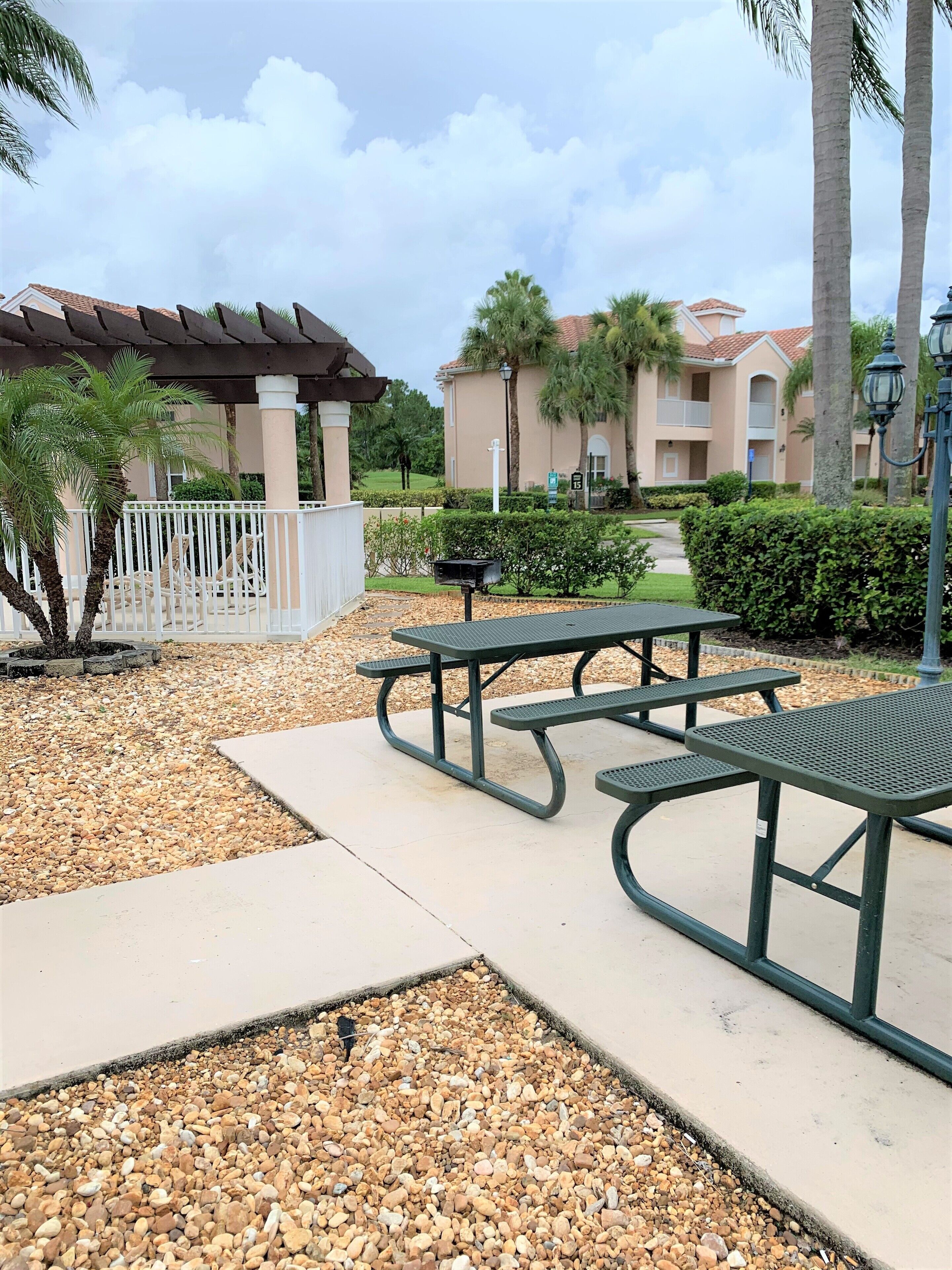 A patio area with picnic tables and a pergola, surrounded by palm trees and a building with a red-tiled roof.