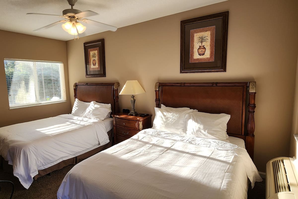 A hotel room with two single beds, a ceiling fan, a window with blinds, and a radiator.