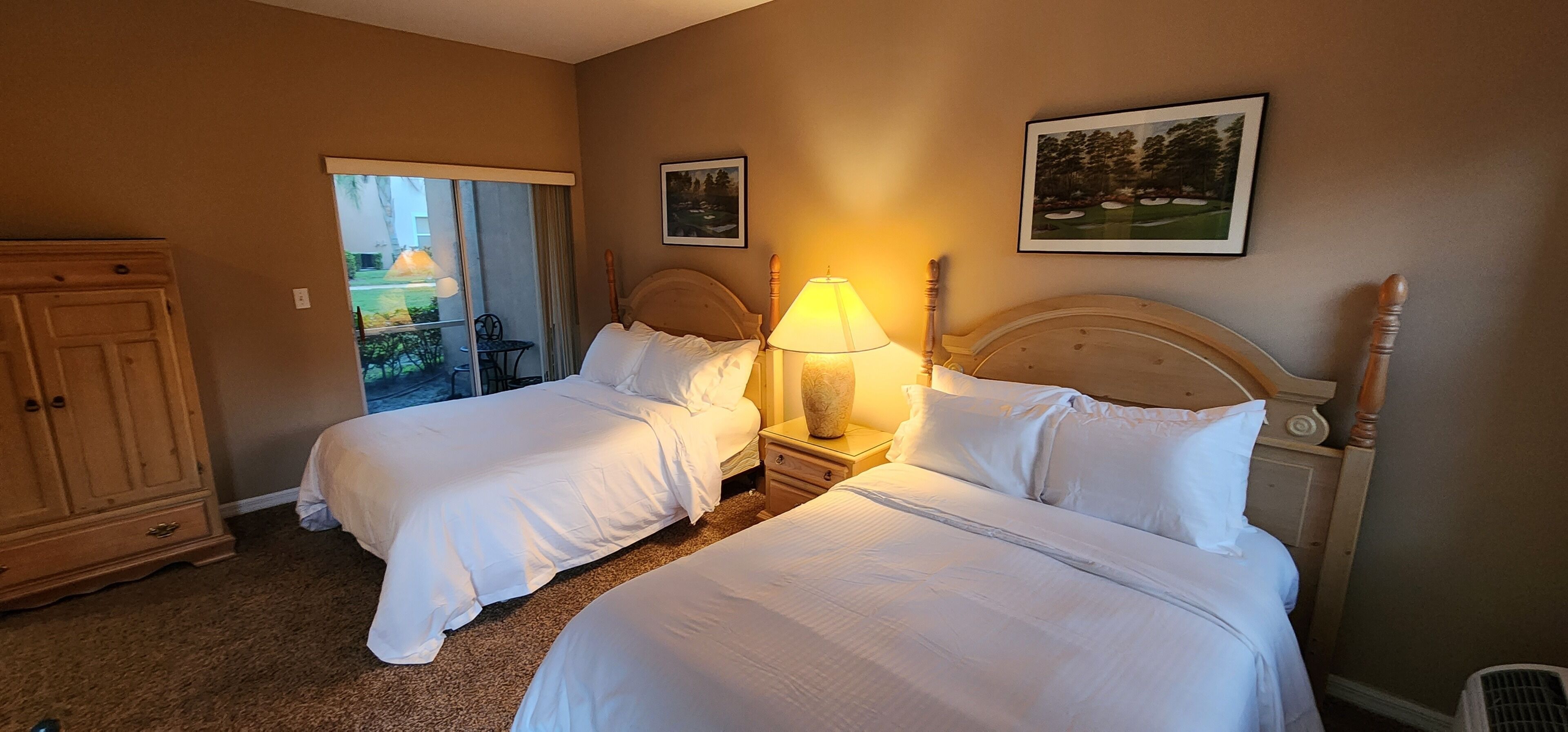 Ein Hotelzimmer mit zwei Betten, einem Nachttisch mit Lampe, einem Holzschrank und einem Balkon mit Tisch und Stühlen.