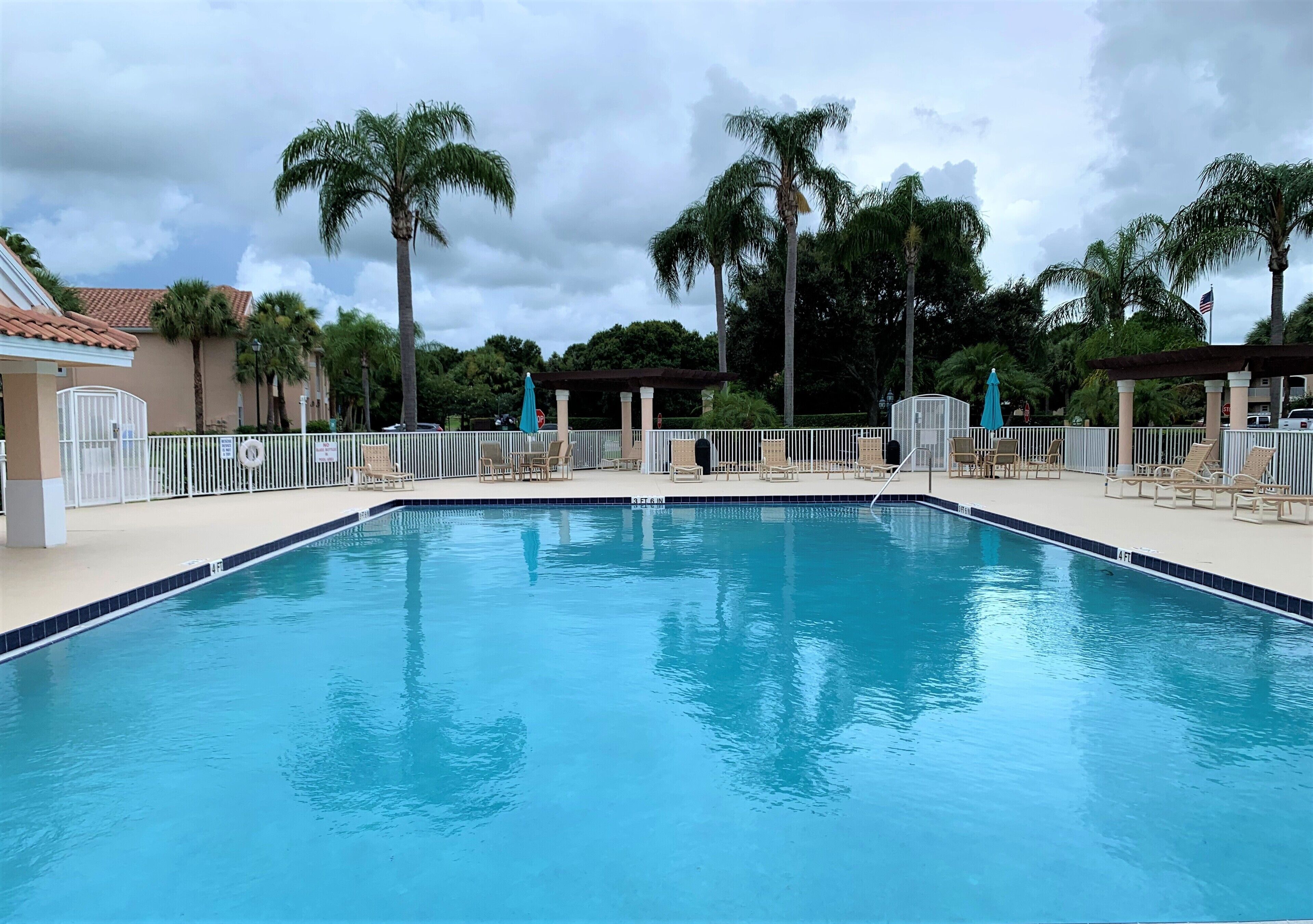 A swimming pool with lounge chairs and umbrellas, surrounded by palm trees and a fence.