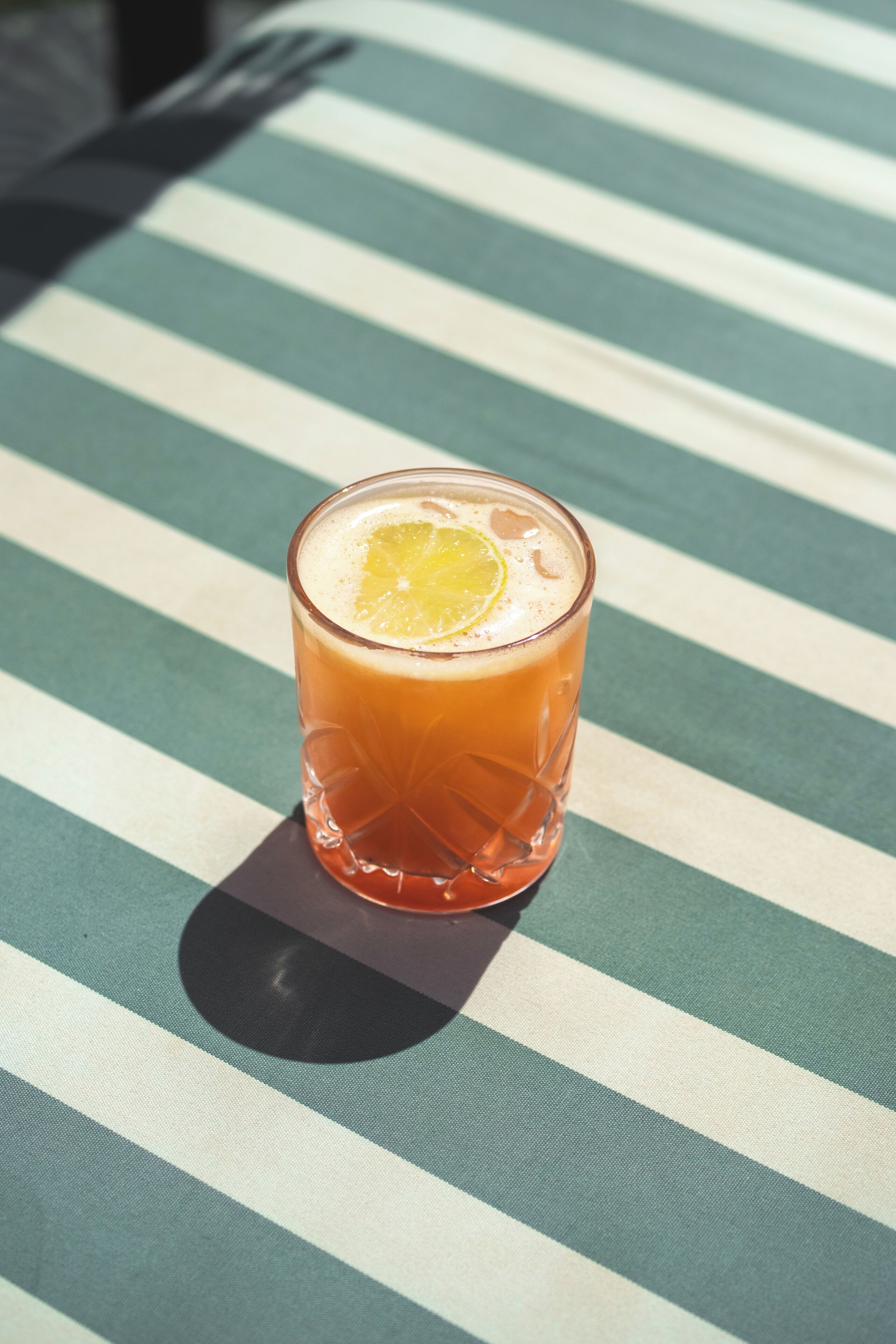 A glass of orange-colored beverage with a slice of orange on the rim, placed on a striped tablecloth.
