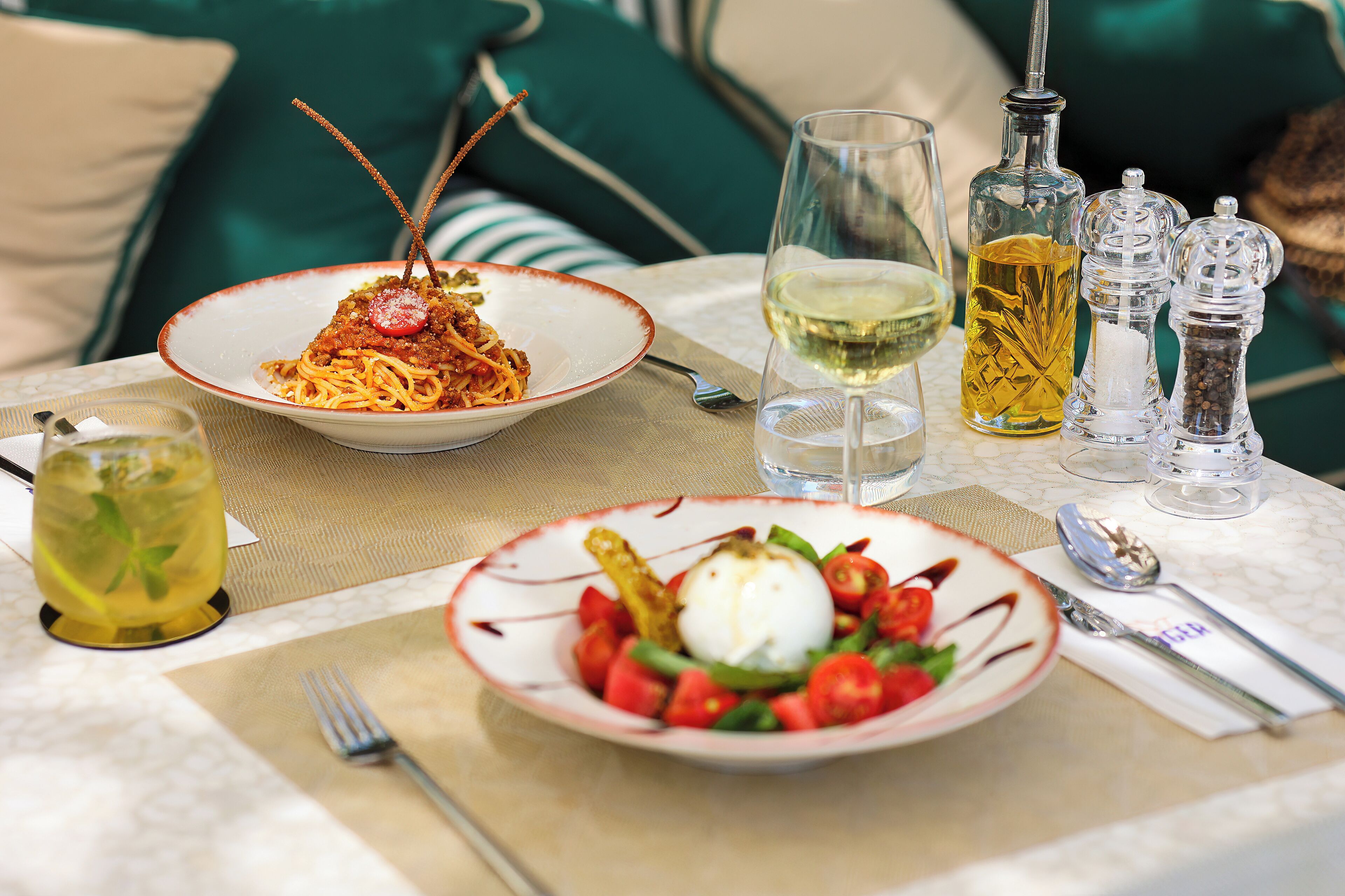 A dining table with two plates of pasta and a salad, a glass of white wine, and a bottle of olive oil.