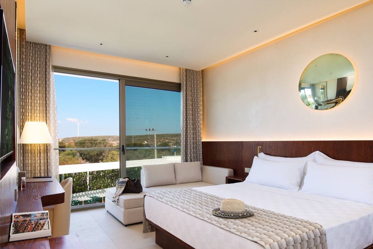A hotel room with a large bed, a desk, a chair, a lamp, and a view of the landscape.