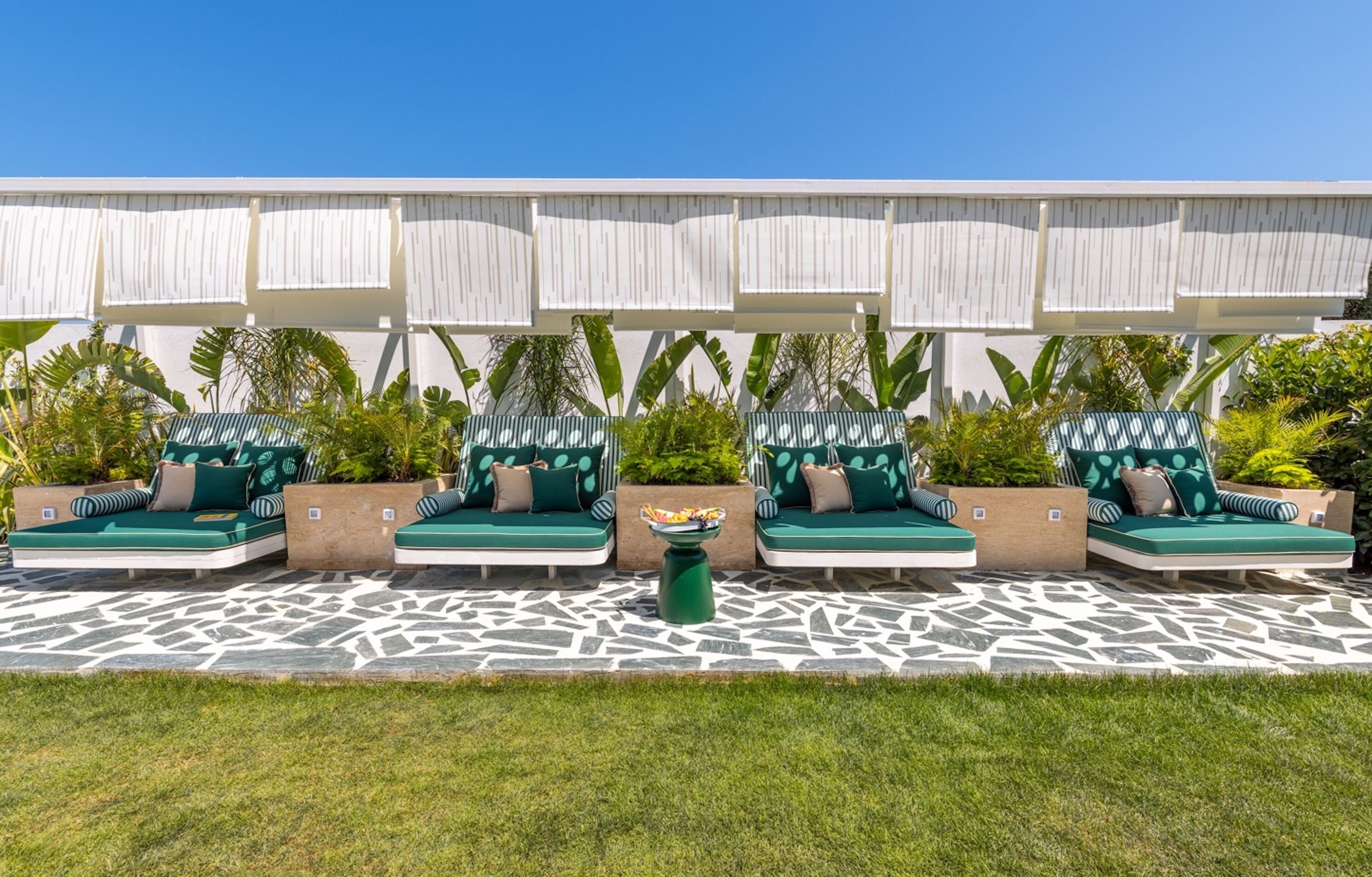 A row of outdoor lounge chairs with green cushions and striped pillows, arranged on a patterned stone patio.