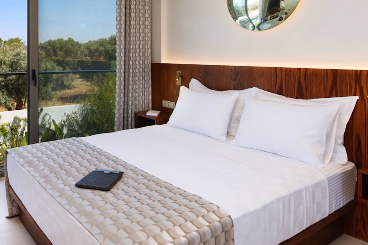 A modern hotel room with a large bed, a round mirror on the wall, and a view of the outdoors through a large window.