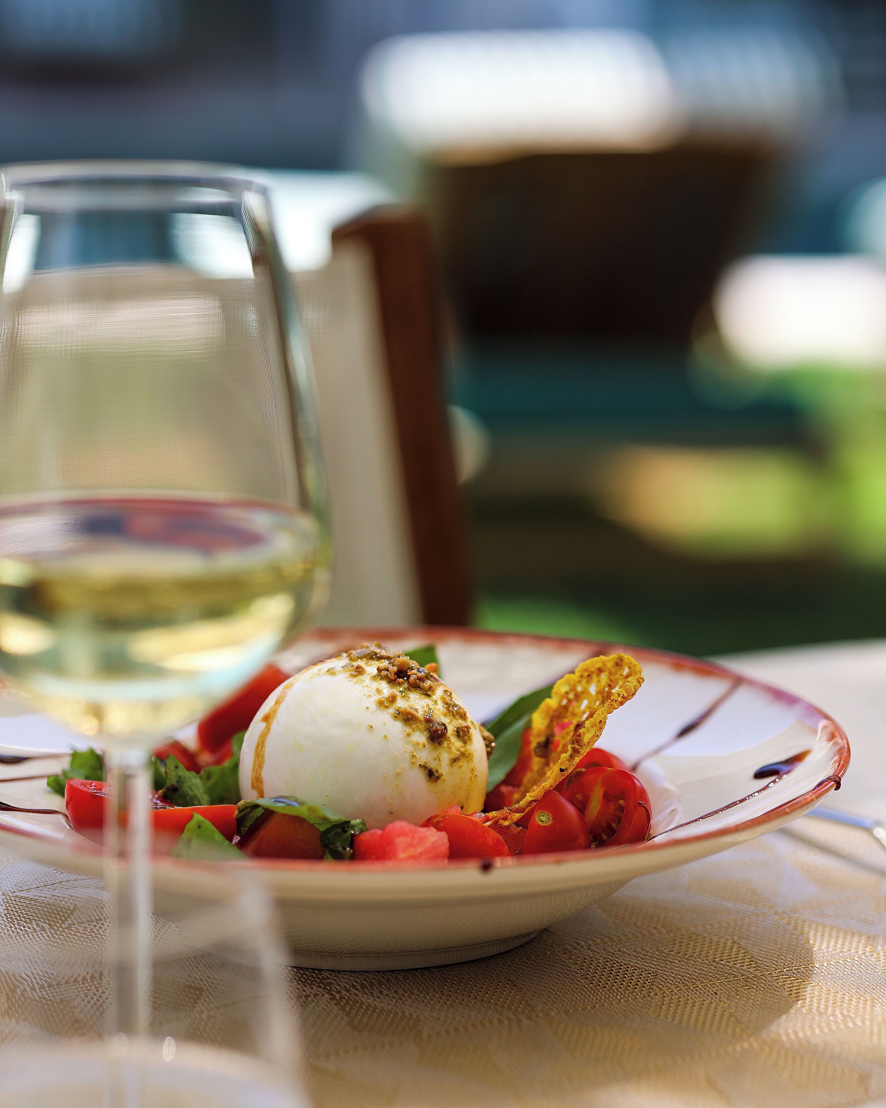 Un plato de comida con una esfera blanca, posiblemente mozzarella, decorado con hierbas y un elemento crujiente, acompañado de tomates cherry y hojas verdes, sobre una mesa con una copa de vino blanco.