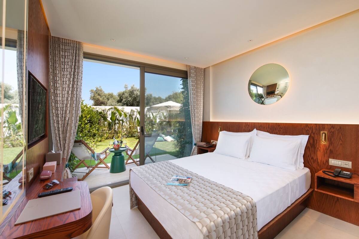 A modern hotel room with a large bed, a desk with a computer, and a view of the outdoors.