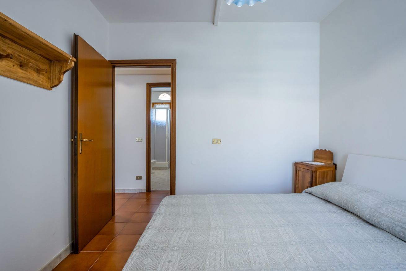 A bedroom with a bed, a wooden nightstand, a door, and a hallway.