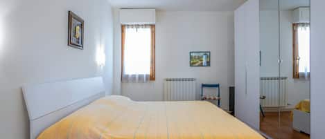 Un dormitorio con cama, un radiador, una ventana con cortinas, un cuadro enmarco y un armario.