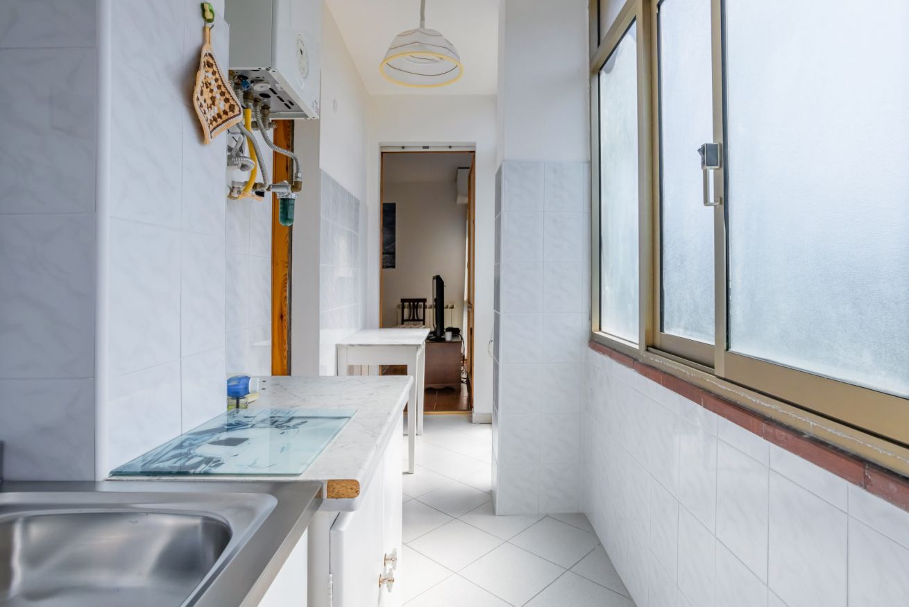 Una cocina estrecha con un ventanal grande, azulejos blancos y un lavamanos de acero inoxidable.
