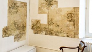 Een kamer met een witte radiator, een houten stoel en twee kunstwerken aan de muur, voorstellende bomen en begroeiing.