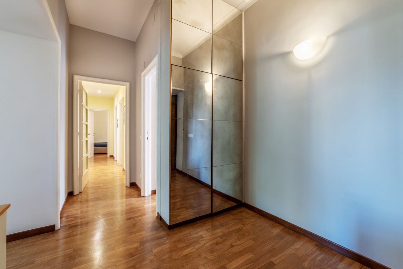Un pasillo con suelo de madera, un muro espejado y una puerta que da a otra habitación.