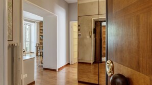 Een gang met houten vloer, een deur met een ronde knop, en zicht op een kamer met raam en stoel.