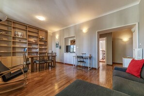 A spacious living area with a wooden floor, a dining table with chairs, a sofa, a bookshelf, and a TV.