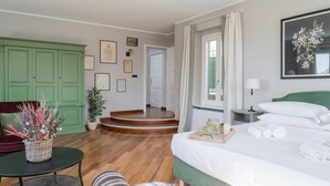 Une chambre d’hôtel comprenant un lit, une table de chevet, une commode, une chaise, une armoire verte, un mur orné de tableaux et un escalier menant à une autre pièce.