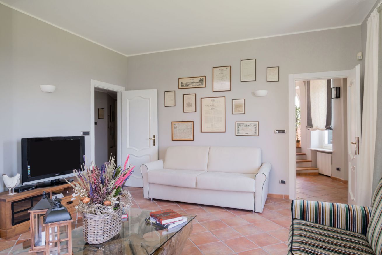 A living room with a television, a white sofa, a coffee table with books and a decorative basket, and framed certificates on the wall.